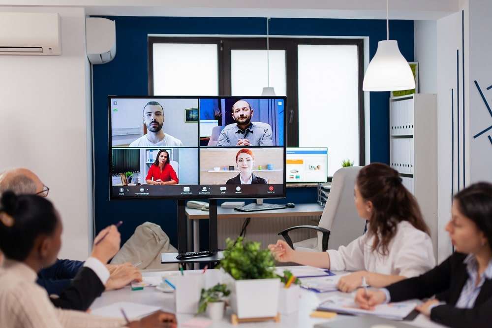 Colleagues around a meeting-room table watching a wall screen showing four people on a video call
