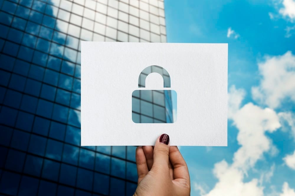A hand holding up a sheet of paper with a padlock cut out of it, against an office tower and blue sky